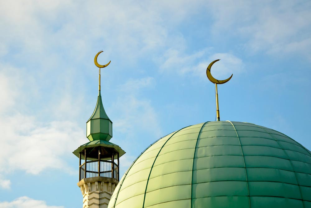 Stor grön kupol och en minaret på en modern moské med varsin gyllene halvmånssymbol mot en blå himmel